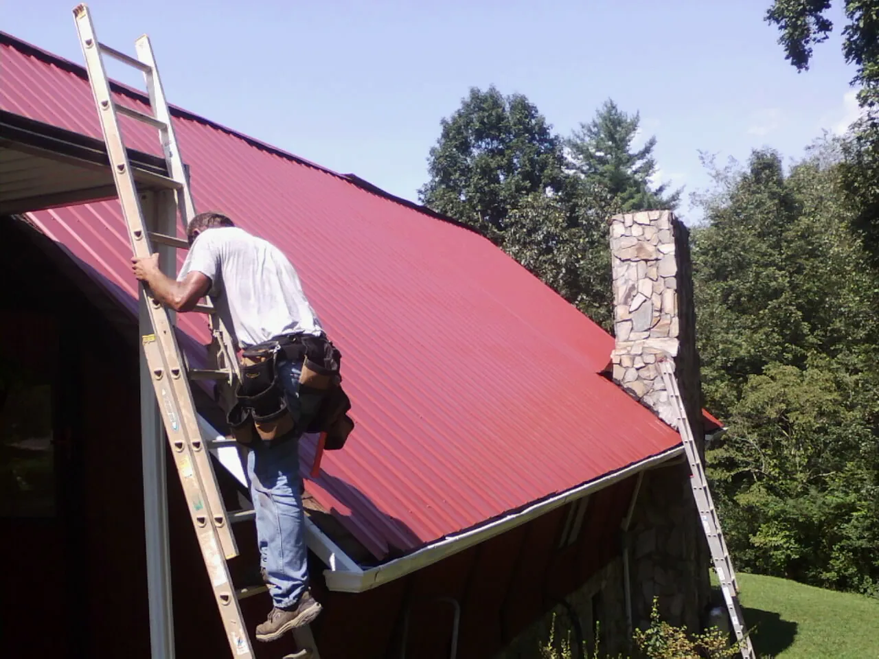 Roofer on a ladder beside a red metal roof and stone chimney — Soffit & Fascia Repair by Roofing Brothers in Covesville